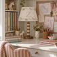 A styled children's desk featuring a decorative table lamp with a cream fabric shade printed with pastel rainbows and a large letter 'Z', alongside a pink-and-white striped chair, books, pencils, and framed unicorn artwork.