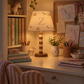 A cozy, warmly lit children's study desk featuring a decorative table lamp with a unicorn-patterned shade and a striped base. The desk is styled with books, colorful pencils, a small unicorn figurine, a vase of daisies, and a pink-and-white striped chair in the foreground.