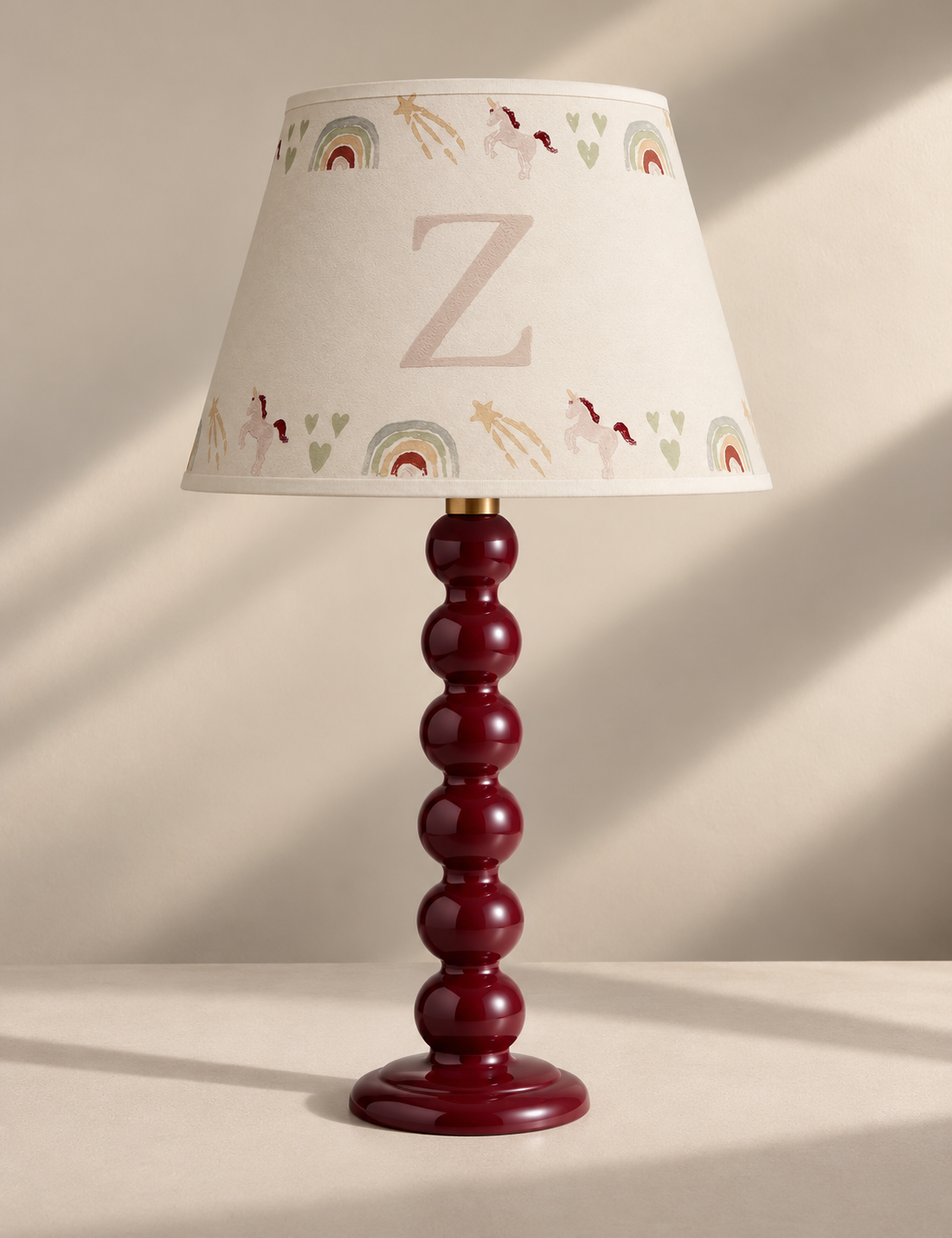 A personalized table lamp with a glossy dark red beaded stem and a cream-colored conical shade decorated with a large letter 'Z', rainbows, unicorns, and hearts.
