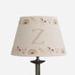 A conical table lamp shade with a beige linen-like fabric printed with pastel rainbow arches, small unicorns, stars, and hearts, featuring a large monogram letter 'Z' in the center. The shade is mounted on a dark metal lamp stand against a plain white background.