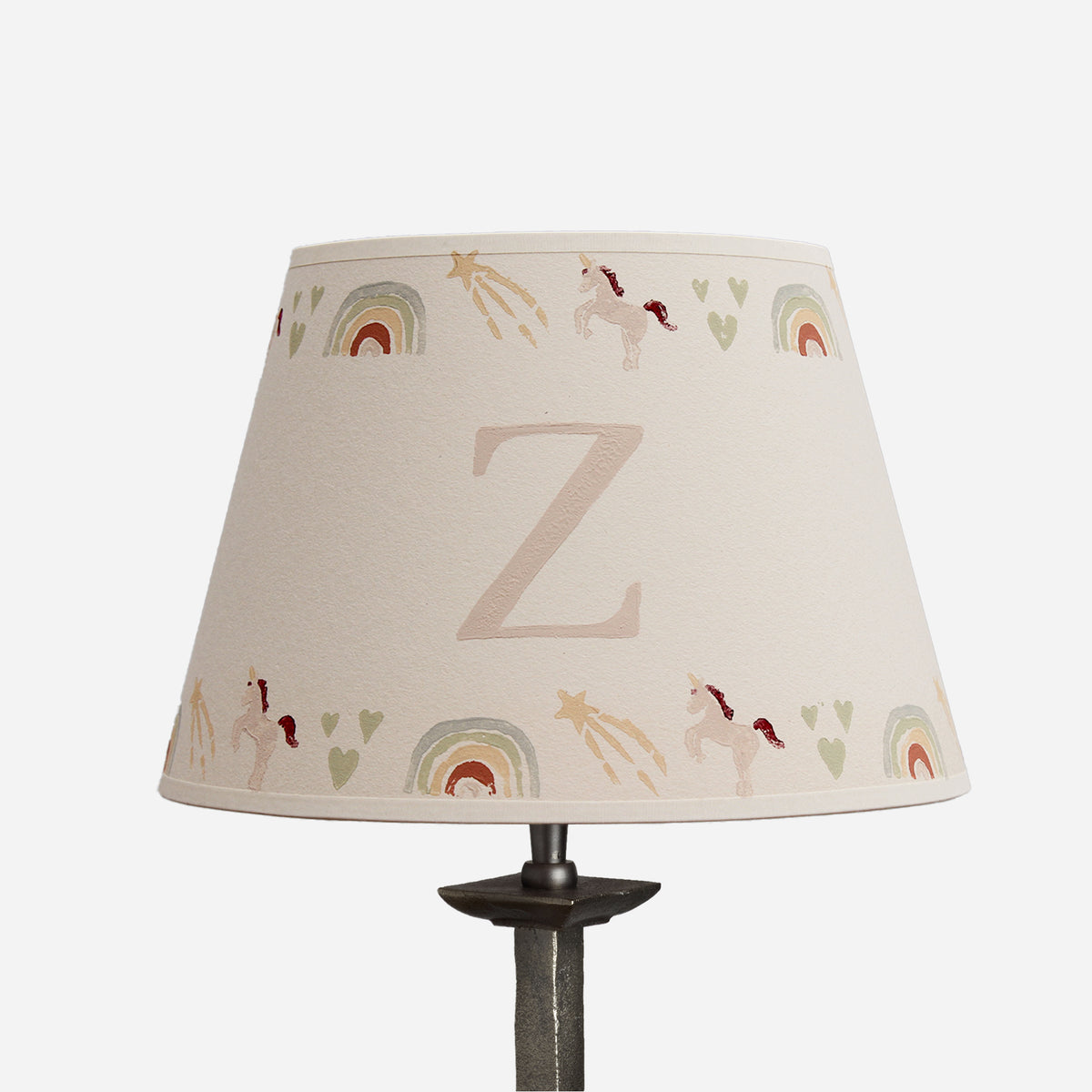 A conical table lamp shade with a beige linen-like fabric printed with pastel rainbow arches, small unicorns, stars, and hearts, featuring a large monogram letter 'Z' in the center. The shade is mounted on a dark metal lamp stand against a plain white background.