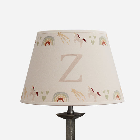A conical table lamp shade with a beige linen-like fabric printed with pastel rainbow arches, small unicorns, stars, and hearts, featuring a large monogram letter 'Z' in the center. The shade is mounted on a dark metal lamp stand against a plain white background.