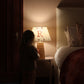 A dimly lit bedroom with a glowing personalised lampshade featuring and a woodland pattern. A young girl stands beside it, reaching for the lamp.