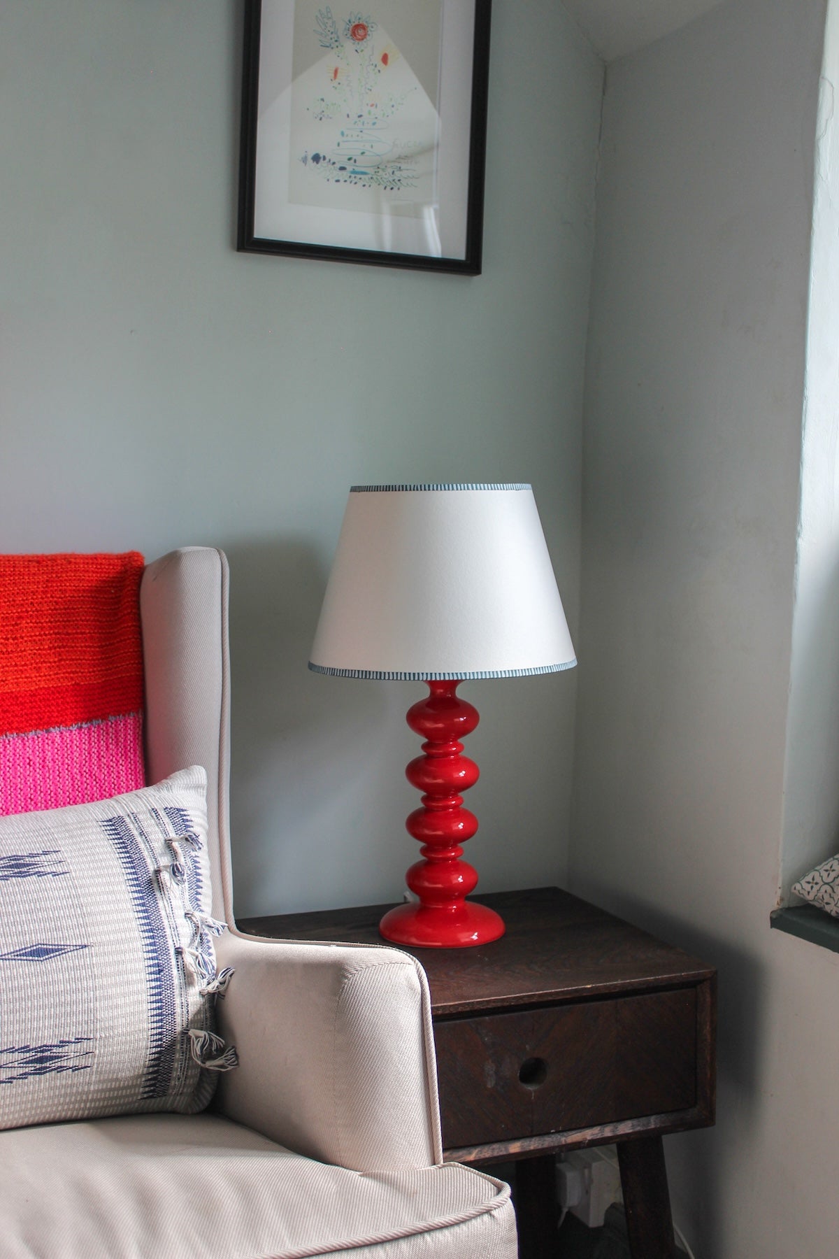 Empire lampshade with hand-painted striped edge in soft blue, styled on a bold red table lamp. A colourful lampshade design adding a touch of striped lighting charm to a modern living room.